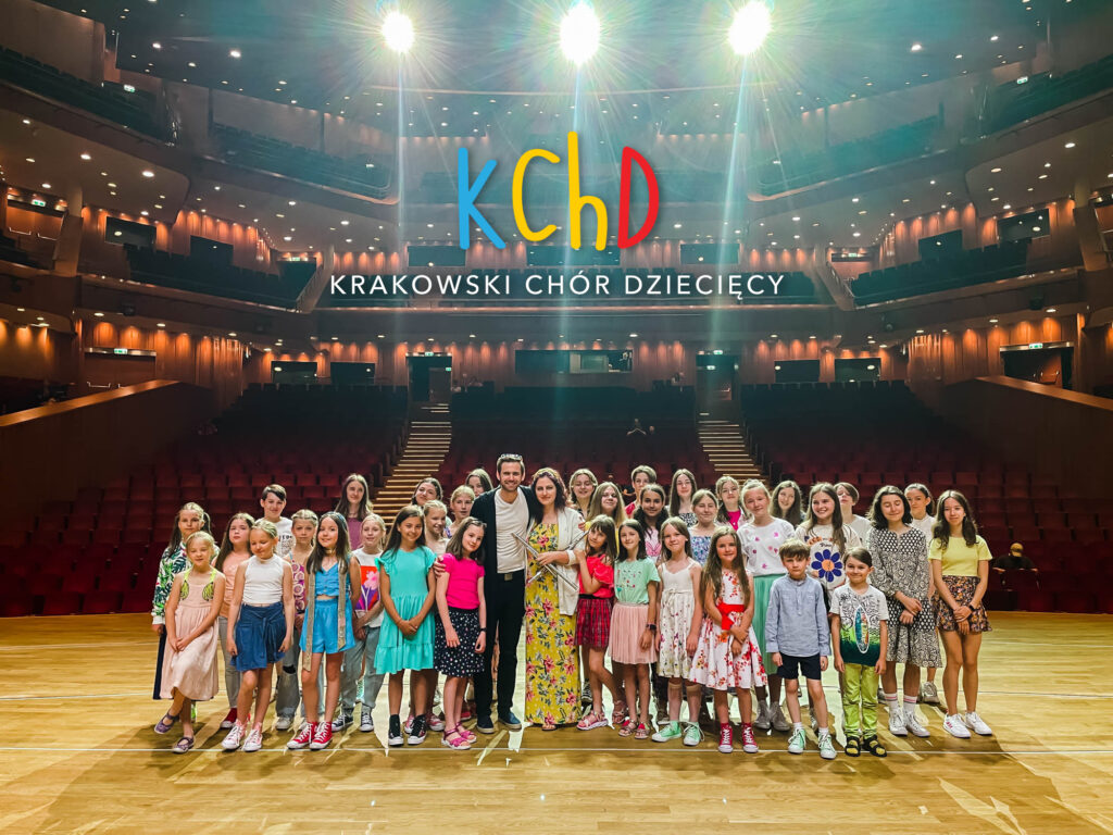 Krakowski chór dzieciecy. Bardzo liczna grupa stoi na oświetlonej scenie, w tle widać wnętrze sali koncertowej